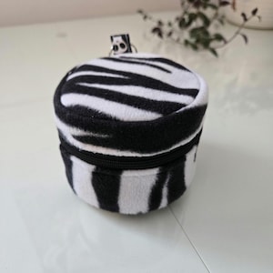 Pode incluir: Pequena bolsa redonda com zíper e padrão de listras de zebra em preto e branco. A bolsa tem um zíper preto e um pequeno chaveiro. Feita de um material macio e felpudo.