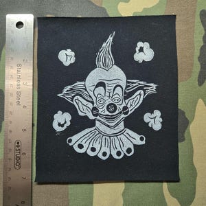 Op de afbeelding: Een vierkante zwarte stoffen patch met een witte zeefdrukillustratie van een clown met een puntig kapsel, een geplooide kraag en vier wolkachtige vormen. Een roestvrijstalen liniaal toont aan dat de patch 20,3 cm hoog is.