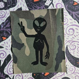 Könnte beinhalten: Ein rechteckiger Stoffaufnäher mit einem Tarnmuster in Grün-, Braun- und Beigetönen. In der Mitte befindet sich die schwarze Silhouette eines Alien-Wesens mit großem Kopf und ovalen Augen, das mit einer Hand ein Friedenszeichen zeigt.