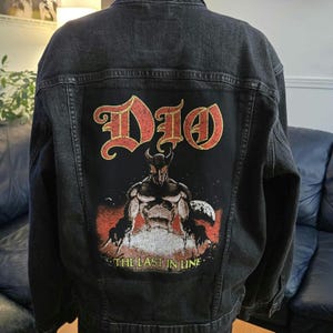 Może przedstawiać: Czarna kurtka dżinsowa z dużą grafiką na plecach. Grafika przedstawia nazwę zespołu "Dio" w czerwonych i złotych literach nad ilustracją rogatej postaci. Tekst "THE LAST IN LINE" znajduje się poniżej.