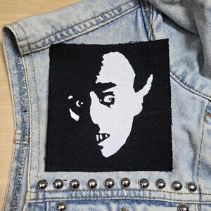 May include: A square black fabric patch featuring a high-contrast white screen-printed graphic of a human face with sharp, pointed ears and visible teeth. The patch is sewn onto a light blue acid-wash denim vest adorned with silver metal studs.