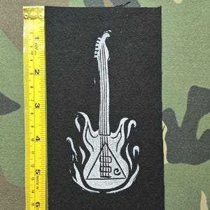 Peut inclure: Un écusson rectangulaire en tissu noir avec un motif blanc représentant une guitare électrique entourée de flammes. Un ruban à mesurer jaune est placé le long du bord gauche, indiquant que l'écusson mesure environ 15 cm de longueur.