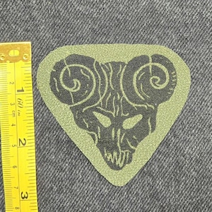 Peut inclure: Un écusson en tissu vert olive de forme triangulaire arborant un motif noir représentant un crâne de bélier aux cornes en spirale. Un mètre ruban jaune est placé à gauche, indiquant une largeur d'environ 7,6 cm et une hauteur de 7 cm.