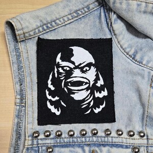 May include: A square black fabric patch featuring a white screen-printed graphic of a creature's face with large eyes and a wide mouth, sewn onto a light blue denim vest adorned with a horizontal row of silver-toned metal studs along the bottom.