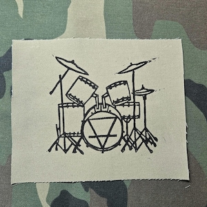 Op de afbeelding: Een rechthoekige stoffen patch in een licht olijfgroene kleur met een zwarte lijntekening van een drumstel. De afbeelding toont een basdrum met een driehoekssymbool erin, twee toms, een floortom en twee bekkens op standaards.