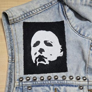 May include: A light blue acid-wash denim waistcoat featuring a rectangular black fabric patch with a white screen-printed graphic of a stylised human face. The waistcoat is accented with a horizontal row of silver-toned metal studs along the lower seam.