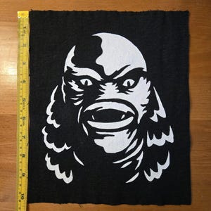 May include: A black fabric patch featuring a white screen-printed graphic of a creature's face with large eyes and a wide mouth. A yellow measuring tape is placed along the left edge, showing the patch is approximately 10 inches in height.
