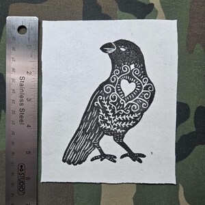 Peut inclure: Un écusson carré en tissu blanc avec une illustration à l'encre noire représentant un oiseau orné de motifs en filigrane et d'un cœur sur la poitrine. Une règle en acier inoxydable indique une hauteur d'environ 15 cm pour l'écusson.