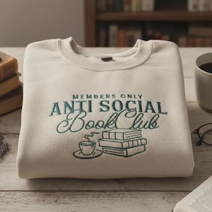 May include: Cream-colored sweatshirt with the text "MEMBERS ONLY ANTI SOCIAL Book Club" embroidered in teal. The design includes a teacup, saucer, and stacked books. The sweatshirt is folded on a wooden surface.