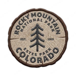 May include: Embroidered patch featuring the text "Rocky Mountain National Park Estes Park Colorado" around a tree graphic. The patch is circular, with a brown border and a light brown, wood-grain-like center.