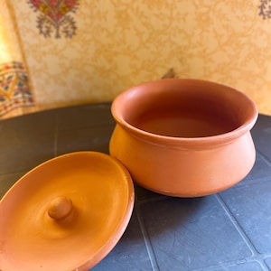 May include: An open, terracotta-colored clay pot with a matching lid. The lid is off to the side, revealing the interior of the pot. The pot has a rounded shape and a simple design, suitable for cooking or serving.