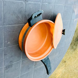Puede incluir: Una olla naranja con tapa, asas negras y borde negro. La olla está abierta, revelando su interior. La olla está sobre una mesa gris.