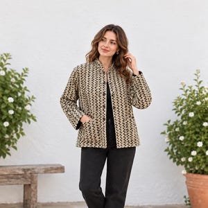 May include: A long-sleeve jacket featuring a repeating black and tan geometric pattern, a mandarin collar, a front button closure, and two large patch pockets on the front, paired with black denim trousers and white trainers.
