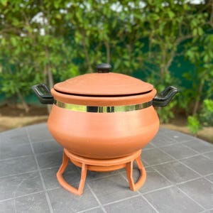 Op de afbeelding: Een terracotta-kleurige aarden pot met een bijpassend deksel en een zwarte handgreep aan de bovenkant. De pot is voorzien van een metallic gouden band in het midden, twee zwarte zijhandgrepen en rust op een bijpassende ronde standaard.