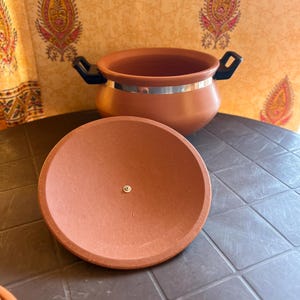 Puede incluir: Una olla de cocina de terracota con su tapa a juego, sobre una superficie oscura. La olla tiene asas negras y una banda plateada alrededor del borde. La tapa es redonda.