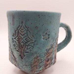 May include: A turquoise ceramic mug with a textured surface featuring a dark brown embossed geometric pattern on the front and a small flower motif. The mug has a speckled finish, a thick handle, and a slightly irregular, hand-formed shape.