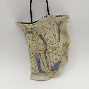 May include: A wall-mounted ceramic vase with an irregular, wavy rim and a cream-colored glaze. The surface features dark blue accents and intricate black calligraphic text. A dark blue braided cord is attached to the top for hanging purposes.
