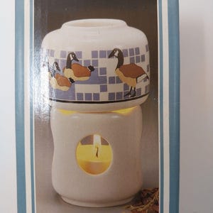 May include: A white ceramic oil warmer featuring a top bowl decorated with a pattern of brown and black geese against a light blue grid background. The base has a circular cutout revealing a lit yellow candle inside the hollow ceramic structure.