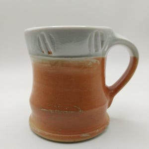 May include: A ceramic mug featuring a two-tone design with a glossy white upper section and a matte terracotta-colored lower body. The white rim includes vertical indentations, and the handle is finished in the same terracotta tone as the base.