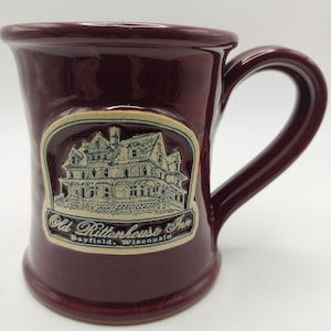 May include: A deep burgundy ceramic mug featuring a raised, cream-colored oval emblem on the front. The emblem displays a detailed illustration of a large house with the text "Old Rittenhouse Inn" and "Bayfield, Wisconsin" printed below it.