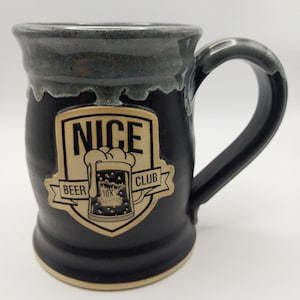 May include: A black ceramic mug with a gray glazed rim and a large handle. The front features a tan shield-shaped emblem with the text NICE BEER CLUB and a graphic of a beer mug with foam. The base of the mug shows a natural tan ceramic finish.