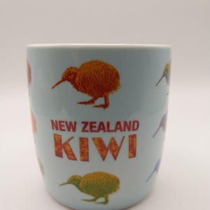 May include: A light blue ceramic mug featuring multiple illustrations of kiwi birds in various colors. The text NEW ZEALAND is printed in red above the word KIWI, which is displayed in a bold, orange, textured font on the front of the mug.