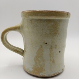 May include: A ceramic mug with a cylindrical body and a curved handle. The exterior features a speckled, off-white glaze with visible brown clay undertones and a darker, textured rim. The surface has an uneven, hand-formed appearance.