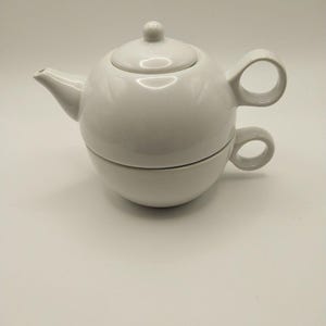 May include: A white ceramic tea set consisting of a teapot stacked on top of a matching teacup. The teapot features a rounded body, a small spout, a lid with a knob, and a circular handle. The teacup has a matching circular handle.