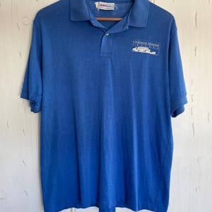 May include: A blue polo shirt with short sleeves, a collar, and a three-button placket. The shirt has the text "Children's Hospital" printed on the front. The shirt is hanging on a wooden hanger.