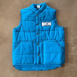 May include: A bright turquoise quilted vest with a stand-up collar and two front pockets. The vest has a button-up closure with brown buttons and a small rectangular patch that reads "HYDRO CONDUIT CORPORATION".