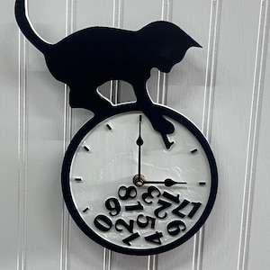May include: A round wall clock featuring a black silhouette of a cat perched on top. The clock face is white with black hands and black numbers that appear to have fallen and gathered at the bottom of the dial. The cat reaches down toward the numbers.