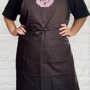 May include: A dark brown apron with a light pink floral design on the chest. The apron has two pockets and a tie at the waist. The person is wearing a black t-shirt and blue jeans.