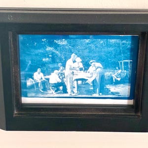 Puede incluir: Fotografía enmarcada en tonos azules que representa a un grupo de personas al aire libre. El marco negro rodea la imagen, que muestra a individuos alrededor de una mesa y sentados en sillas, sugiriendo una reunión social.