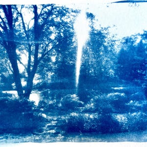 May include: A cyanotype print of a landscape scene. The image features a bright, vertical light source in the center, surrounded by trees and foliage. The print has a vintage, artistic aesthetic, with a blue and white color palette.