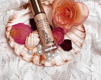 May include: A glass roller bottle with a gold cap and a beige label featuring a mermaid illustration and the text Sacred Spirit Shop Siren's Call Attraction Collection, resting on a large scallop shell with a peach rose, pink petals, and pearls.