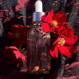 May include: A clear glass bottle with a silver dropper top, filled with a reddish liquid and botanical elements. The bottle is surrounded by vibrant red roses against a black and red lace background.