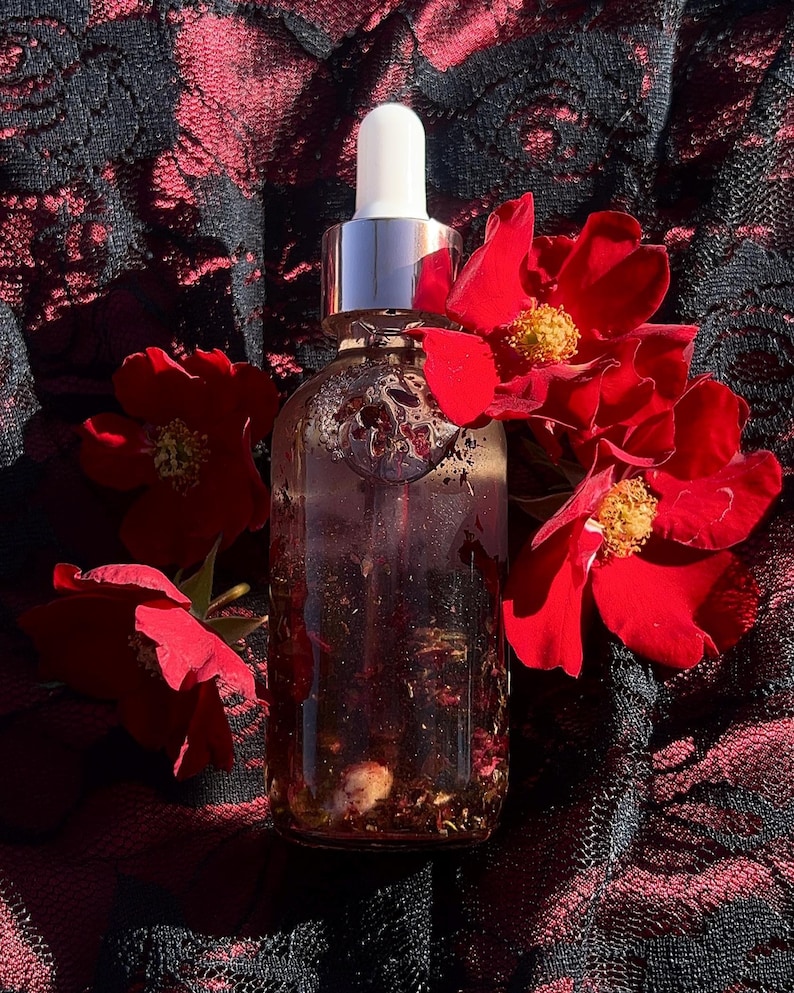 May include: A clear glass bottle with a silver dropper top, filled with a reddish liquid and botanical elements. The bottle is surrounded by vibrant red roses against a black and red lace background.
