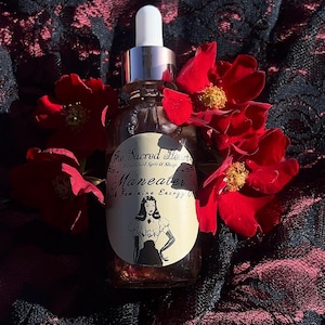 May include: A clear glass bottle with a silver dropper and white cap, labeled "Maneater" with a vintage illustration of a woman. The bottle is surrounded by red roses, set against a dark, patterned background.