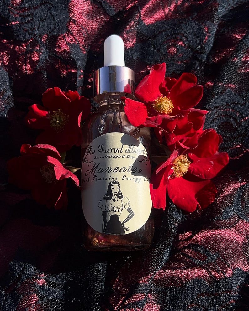 May include: A clear glass bottle with a silver dropper and white cap, labeled "Maneater" with a vintage illustration of a woman. The bottle is surrounded by red roses, set against a dark, patterned background.
