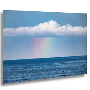 Op de afbeelding: Een foto van een regenboog boven het water. De lucht is lichtblauw met een grote witte wolk. De regenboog is zichtbaar in het midden van de afbeelding, met het water op de voorgrond.