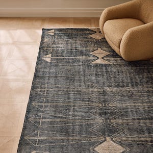 Op de afbeelding: Een donkerblauw rechthoekig vloerkleed met een geometrisch patroon van pijlen en ruitvormen in beige. Het kleed heeft een verweerde uitstraling. Een beige fauteuil is gedeeltelijk zichtbaar in de rechterbovenhoek.