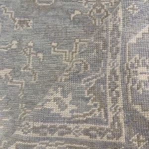 May include: A close-up of a light blue rug with a detailed, repeating pattern in off-white and gray. The rug's texture is visible, with a folded corner revealing the underside. The design features floral and geometric elements.