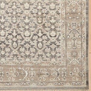 May include: Close-up of a rectangular area rug with a detailed, geometric pattern. The rug features a combination of gray, beige, and off-white colors. The border has a lighter shade of beige with a fringe along the edge.