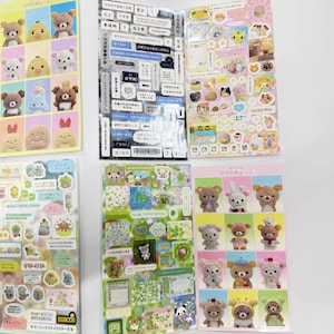 May include: A collection of five sticker sheets featuring various cartoon characters, including Rilakkuma bears and small creatures. The sheets display diverse designs such as grid-based character portraits, text-heavy labels, and assorted decorative icons in multiple colors.