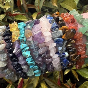 May include: A collection of gemstone chip bracelets in various colors, including green, red, orange, blue, purple, and clear. The bracelets are made of small, irregularly shaped stones strung together. The background features green foliage and a pink surface.