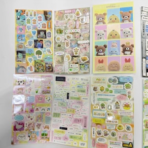 May include: A collection of six rectangular sticker sheets featuring various cartoon characters, including Rilakkuma bears, small birds, and assorted food items. The sheets display colorful illustrations, text labels, and decorative icons in a grid layout.