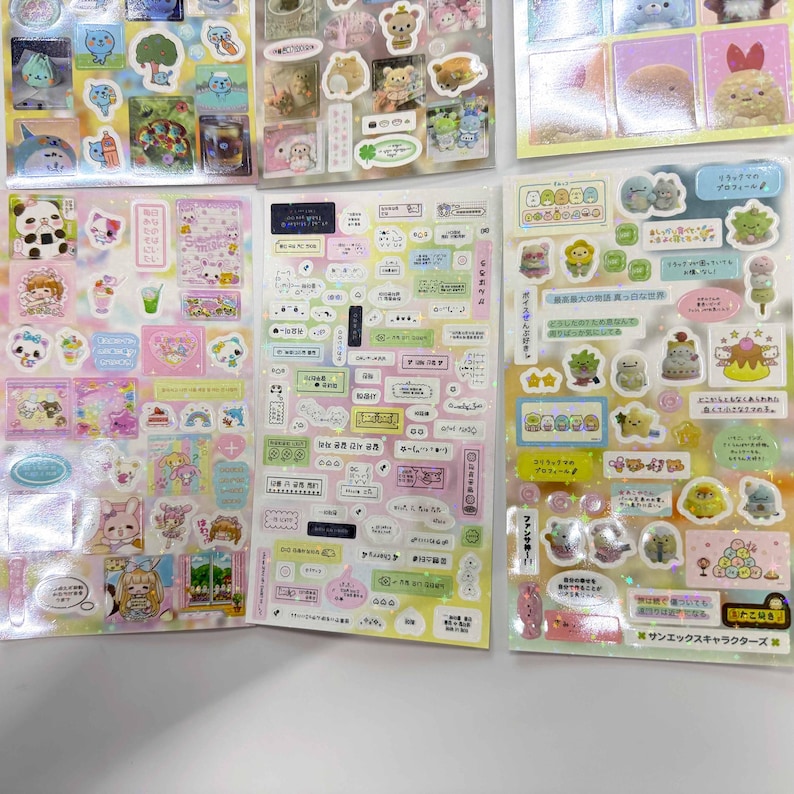 May include: A collection of five sticker sheets featuring various colorful cartoon characters, including bears, cats, and food items, alongside numerous small rectangular labels with Japanese text, all arranged on a plain white surface.