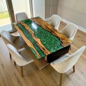 Puede incluir: Una mesa de comedor rectangular con un tablero de madera de borde natural y un río central de resina verde esmeralda translúcida y arremolinada. La mesa se apoya sobre una base oscura y está rodeada por seis sillas tapizadas en tela beige.