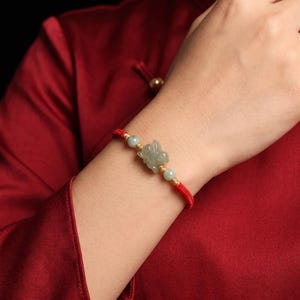 Könnte beinhalten: Ein rotes Kordelarmband mit einem zentralen, geschnitzten hellgrünen Jade-Hasenanhänger, flankiert von zwei kleinen hellgrünen Jadeperlen und goldfarbenen Akzenten. Das Armband wird mit einem einfachen Knotenverschluss befestigt und hat ein rotes Band.
