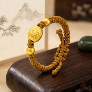 May include: Gold-colored bracelet featuring a central oval charm with a floral design, flanked by two gold spheres. The bracelet is made of a woven, mustard-colored cord. The bracelet is displayed on a dark wooden surface.